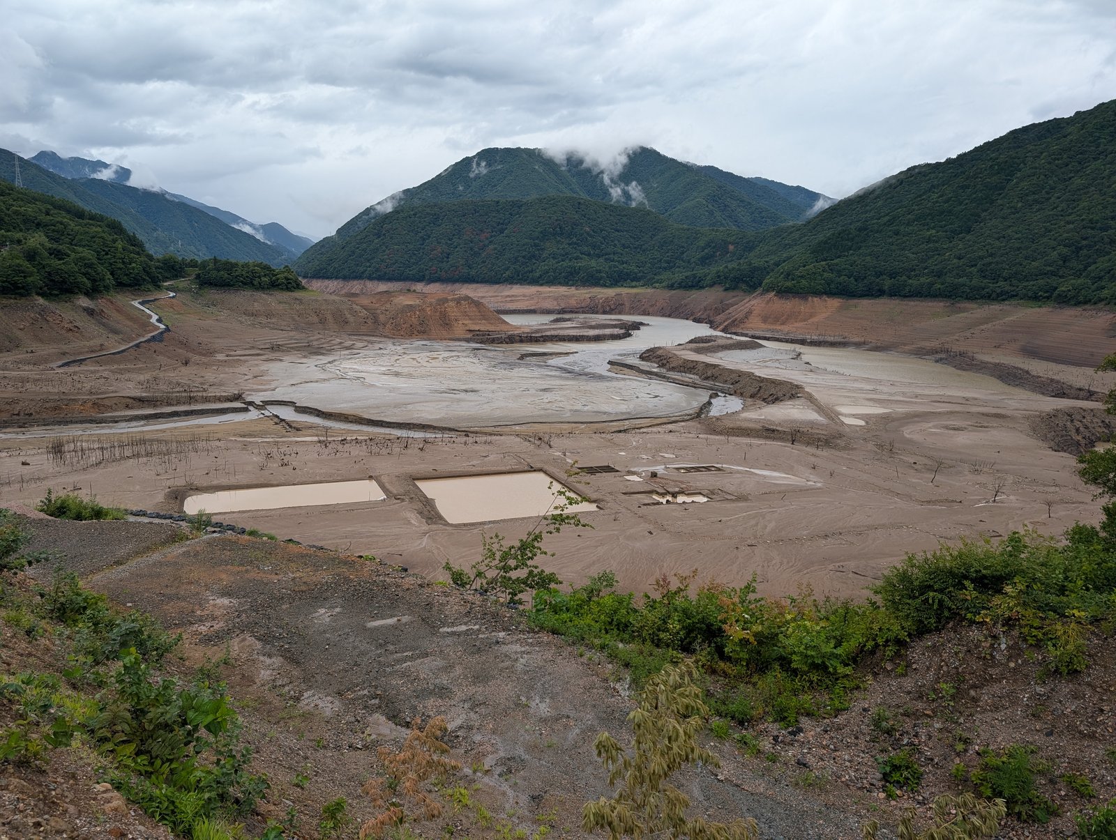 水の引いた御母衣湖。平らな砂泥地に四角い区画がいくつも見え、立ち枯れの木が点々と残る（2026-08-27 撮影）